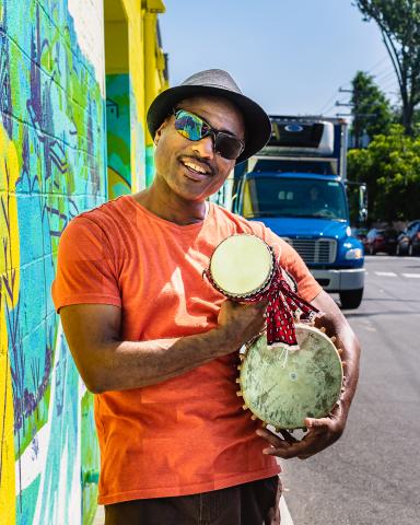 Groovy Nate in a hat and sunglasses outside next to a colorful mural holding an assortment of percussive instruments.